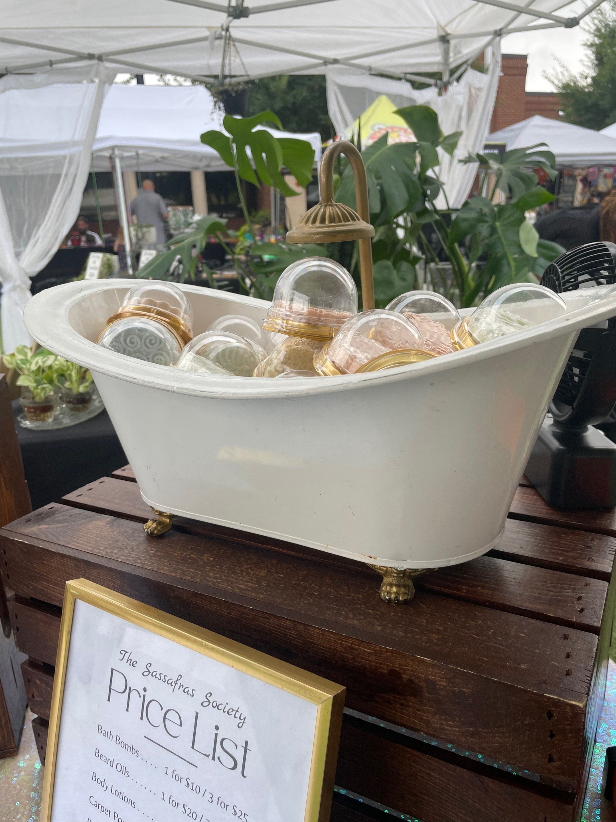 a mini decorative bath tub used to display colorful shower steamers and bathbombs on a wooden crate with plants, people, and other vendor tents in the background